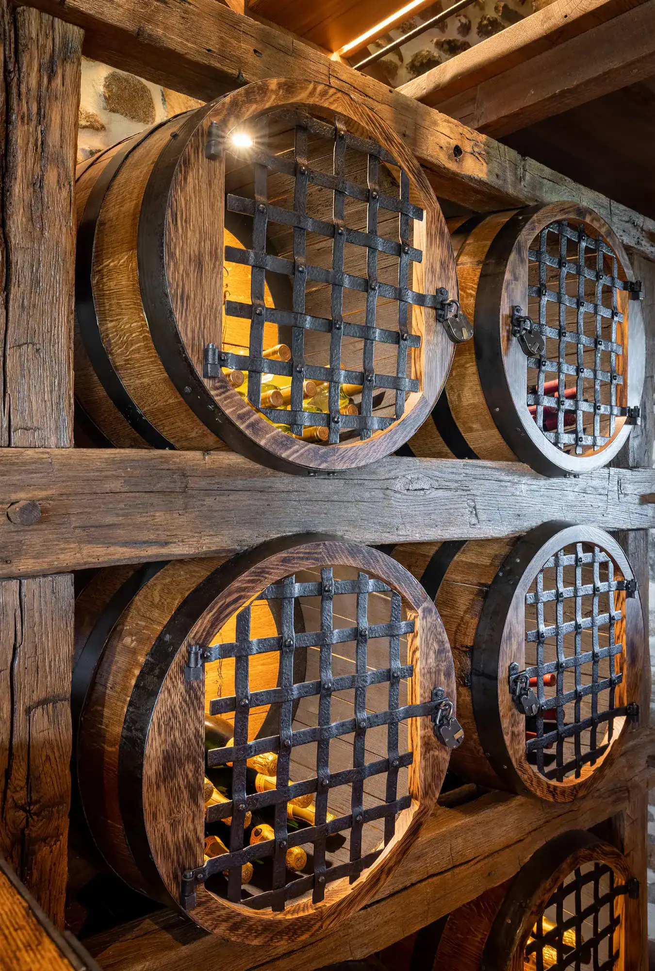 Large wooden barrels with metal grates are built into a rustic wooden wall, storing and displaying wine bottles inside a warmly lit wine cellar reminiscent of a luxury home or party barn.