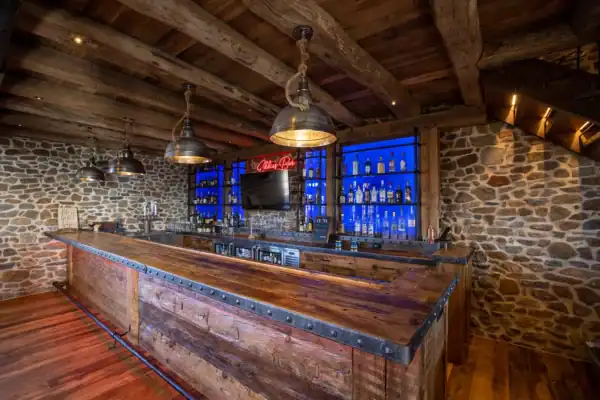 A rustic party barn with a wooden counter and ceiling, stone walls, and industrial-style hanging lights. Behind the bar, shelves display liquor bottles with blue lighting, and a red neon sign reads Lite Beer.
