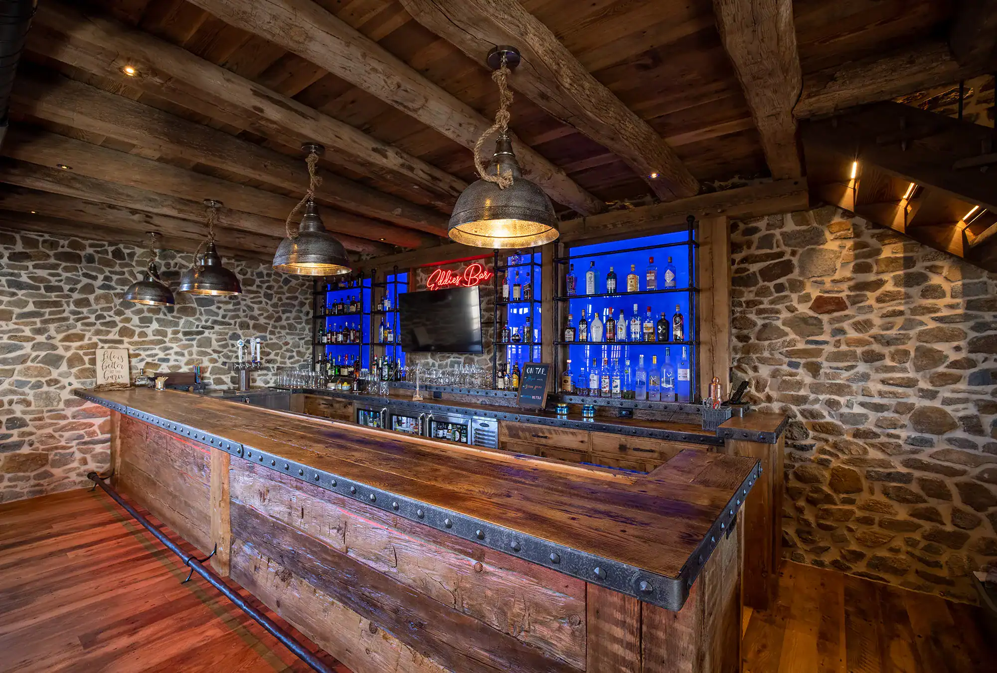 A rustic party barn with a wooden counter and ceiling, stone walls, and industrial-style hanging lights. Behind the bar, shelves display liquor bottles with blue lighting, and a red neon sign reads Lite Beer.