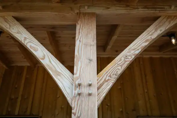 Close-up of timber framing in a luxury horse barn, showing a vertical wooden post joined with diagonal braces using wooden pegs, beneath a wooden roof structure with exposed beams and a hanging light fixture in the background.