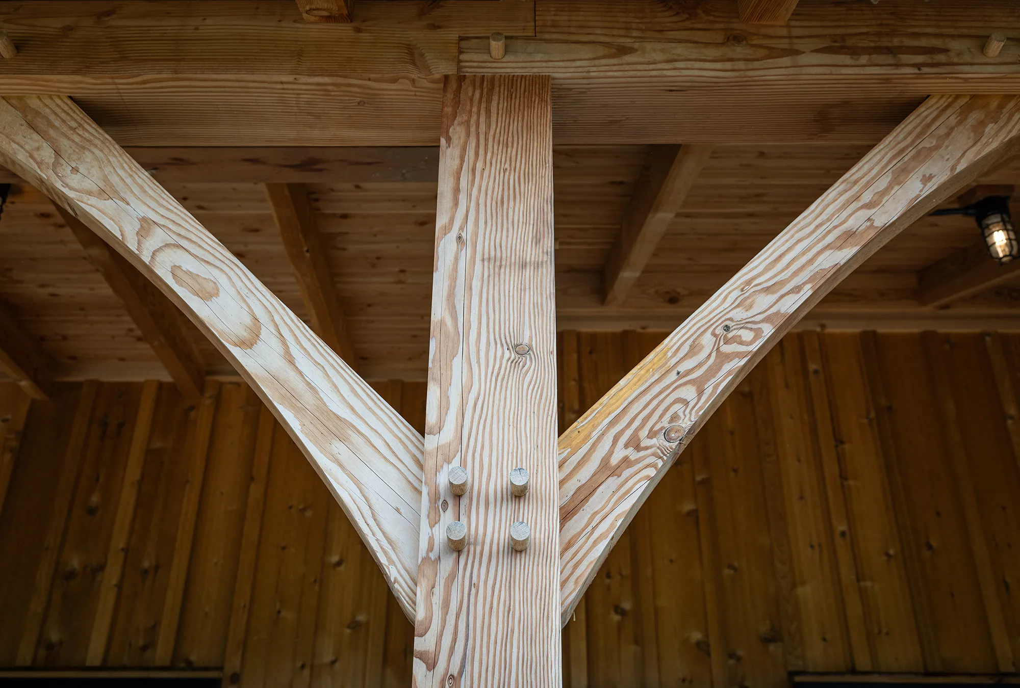 Close-up of timber framing in a luxury horse barn, showing a vertical wooden post joined with diagonal braces using wooden pegs, beneath a wooden roof structure with exposed beams and a hanging light fixture in the background.