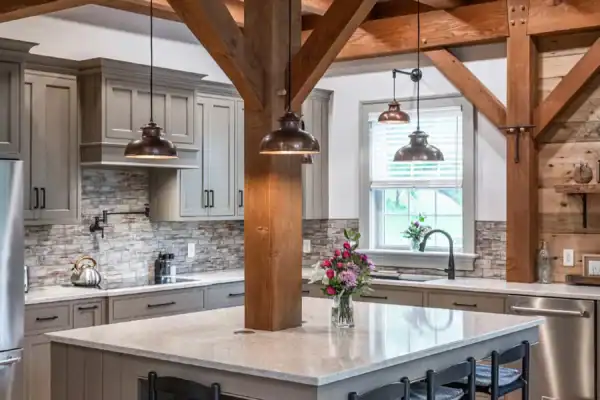 A modern rustic kitchen reminiscent of a luxury home features exposed wooden beams, gray cabinets, stainless steel appliances, a large island with marble countertop, four black stools, pendant lights, and a vase of flowers on the island.