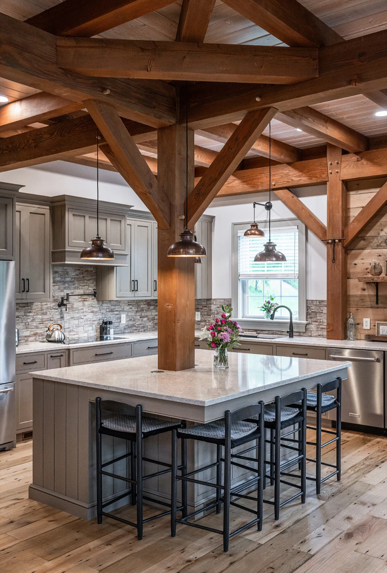A modern rustic kitchen reminiscent of a luxury home features exposed wooden beams, gray cabinets, stainless steel appliances, a large island with marble countertop, four black stools, pendant lights, and a vase of flowers on the island.