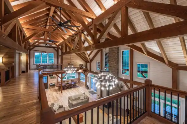 Spacious rustic living area with exposed wooden beams, vaulted ceilings, chandeliers, a stone fireplace, large windows, and a lofted walkway—perfect for gatherings in this elegantly repurposed party barn.