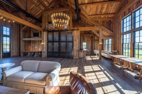 Spacious rustic bank barn room with tall wooden beams, large windows letting in natural light, a sofa and leather chair, round tables, chandeliers, and wood-paneled walls and floor.