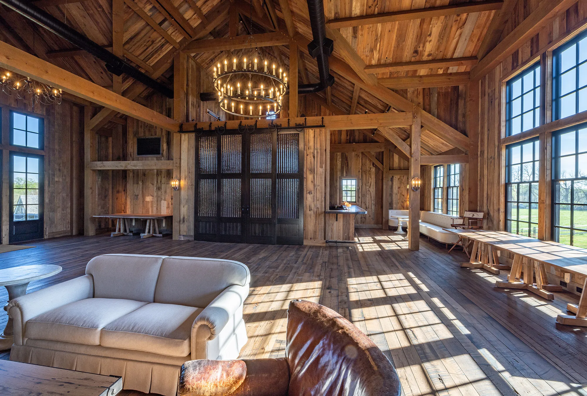 Spacious rustic bank barn room with tall wooden beams, large windows letting in natural light, a sofa and leather chair, round tables, chandeliers, and wood-paneled walls and floor.