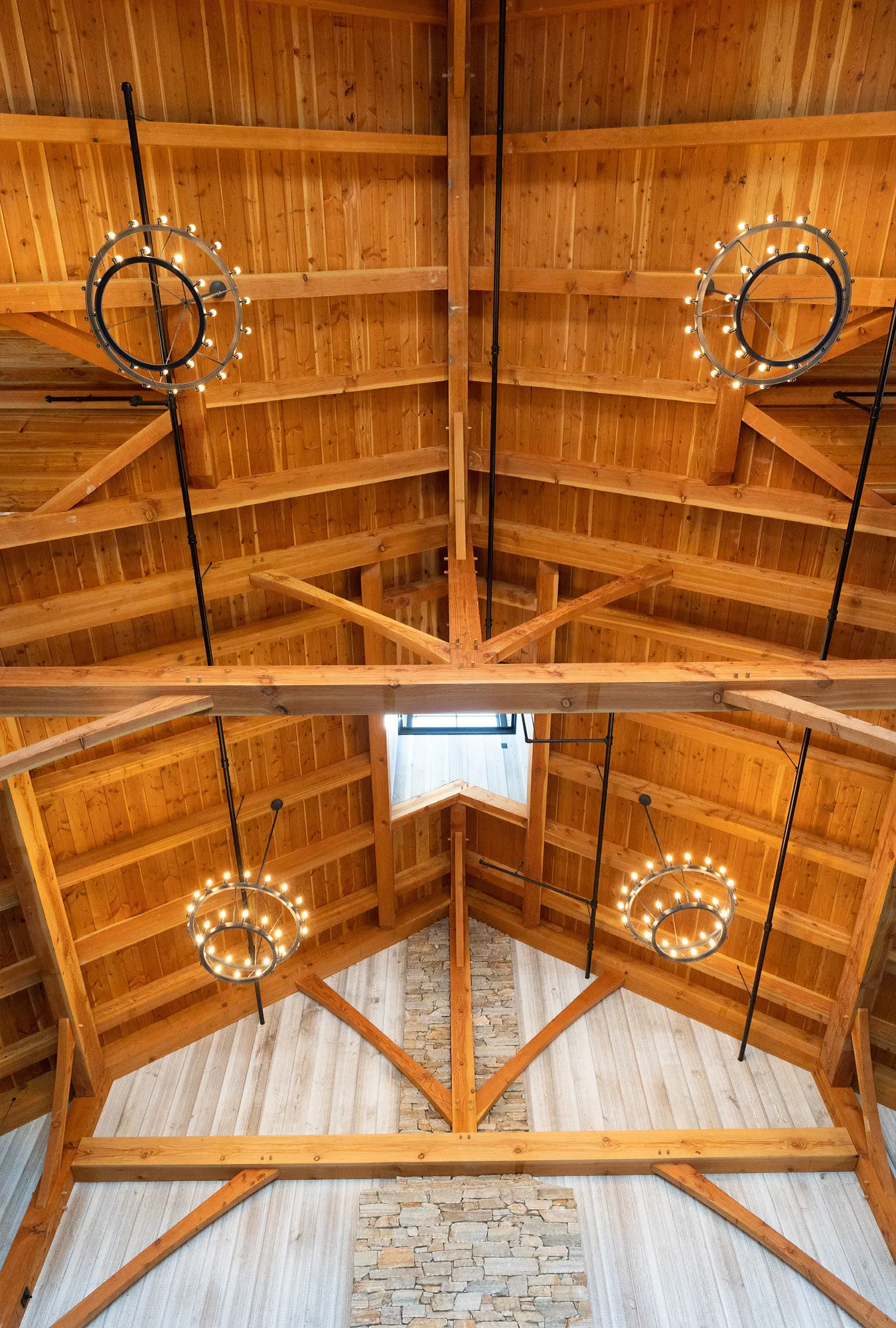 Wooden vaulted ceiling with exposed beams, three circular chandeliers with small lights, and a stone accent wall below a window letting in natural light create the perfect setting for a luxury party barn.