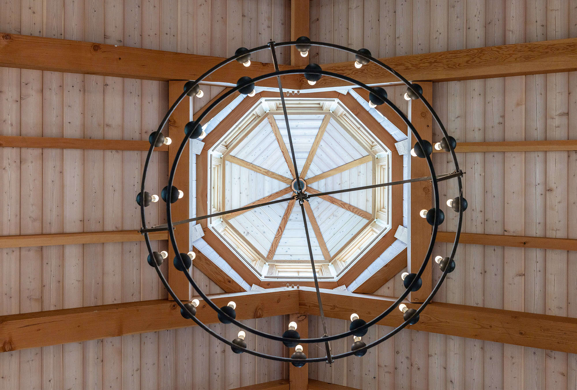 A large circular chandelier with exposed bulbs hangs from a wooden, octagonal vaulted ceiling with visible beams, viewed from directly below—a striking feature in this luxury home. Natural light enters through the central skylight.