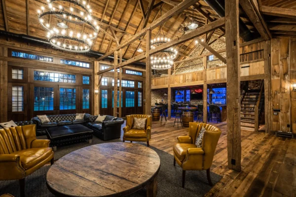 A spacious, rustic lounge in a converted bank barn with exposed wooden beams, leather armchairs, sofas, a round wooden table, large windows, chandeliers, and a bar area with illuminated shelves in the background.