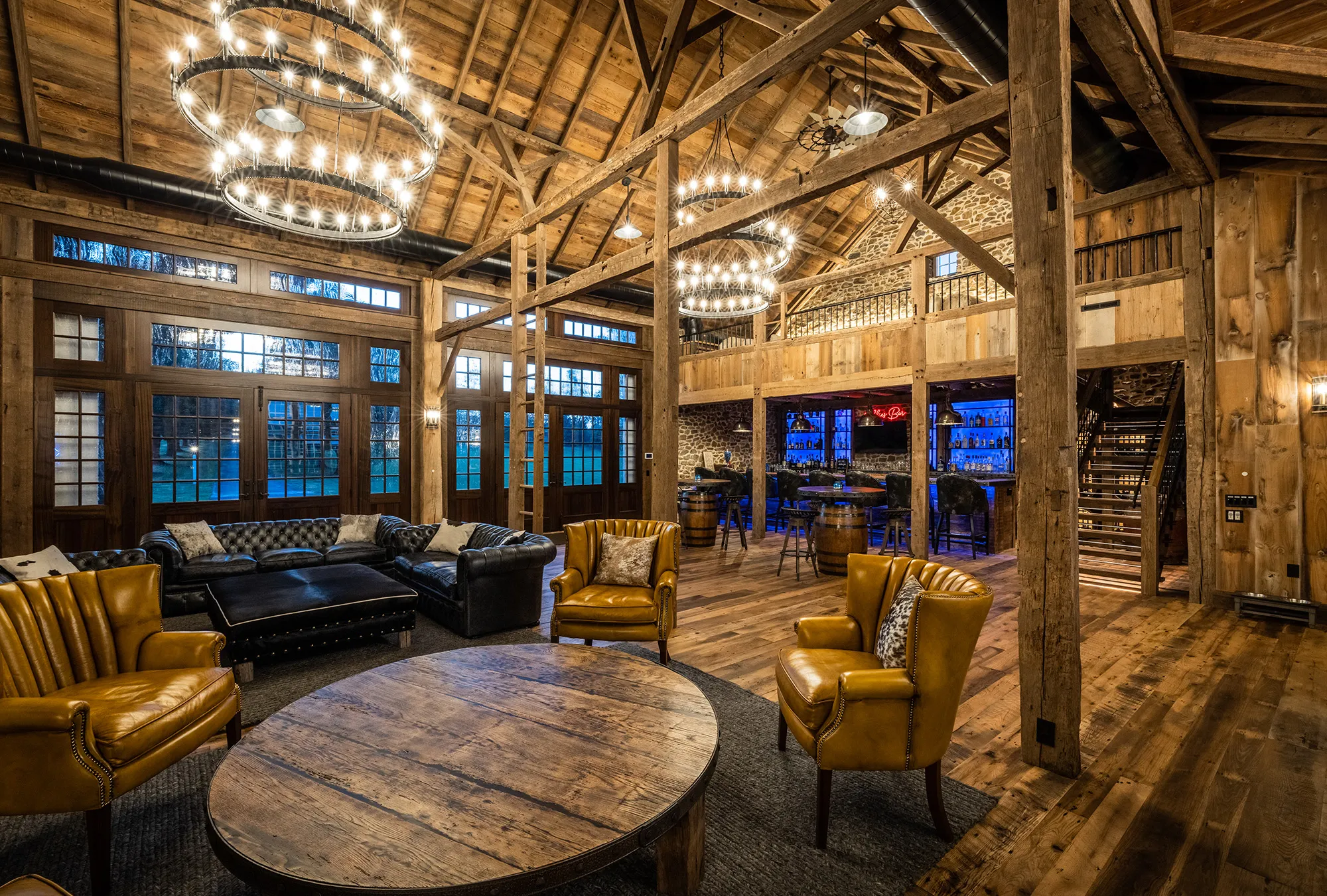 A spacious, rustic lounge in a converted bank barn with exposed wooden beams, leather armchairs, sofas, a round wooden table, large windows, chandeliers, and a bar area with illuminated shelves in the background.