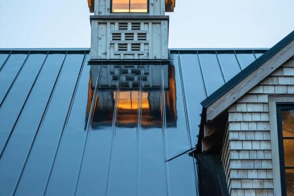 A close-up of a shiny metal roof at dusk reflects light from a windowed cupola above. Warm yellow light glows from the cupola and a nearby house, adding inviting charm to this elegant luxury horse barn against the cool blue evening sky.