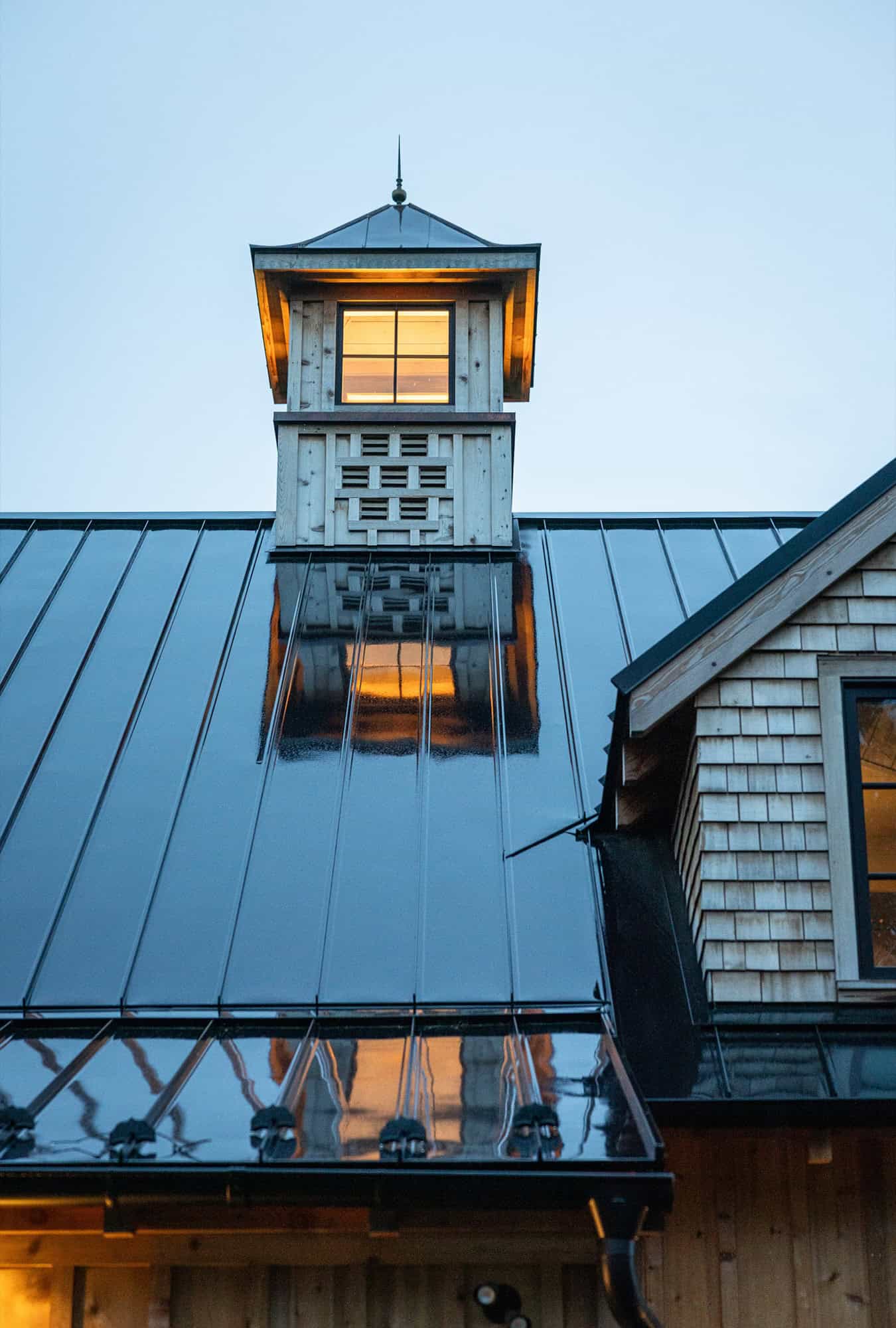 A close-up of a shiny metal roof at dusk reflects light from a windowed cupola above. Warm yellow light glows from the cupola and a nearby house, adding inviting charm to this elegant luxury horse barn against the cool blue evening sky.