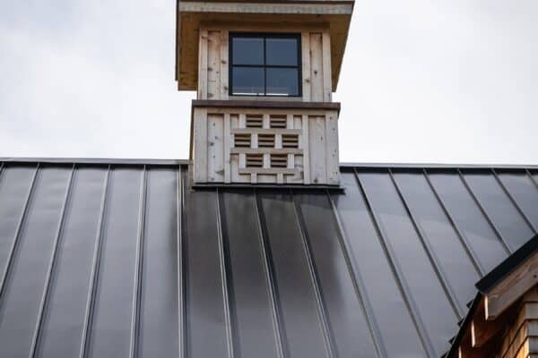 A close-up view of a metal roof with vertical seams on a luxury horse barn, featuring a small wooden cupola with a window and decorative latticework under a cloudy sky.