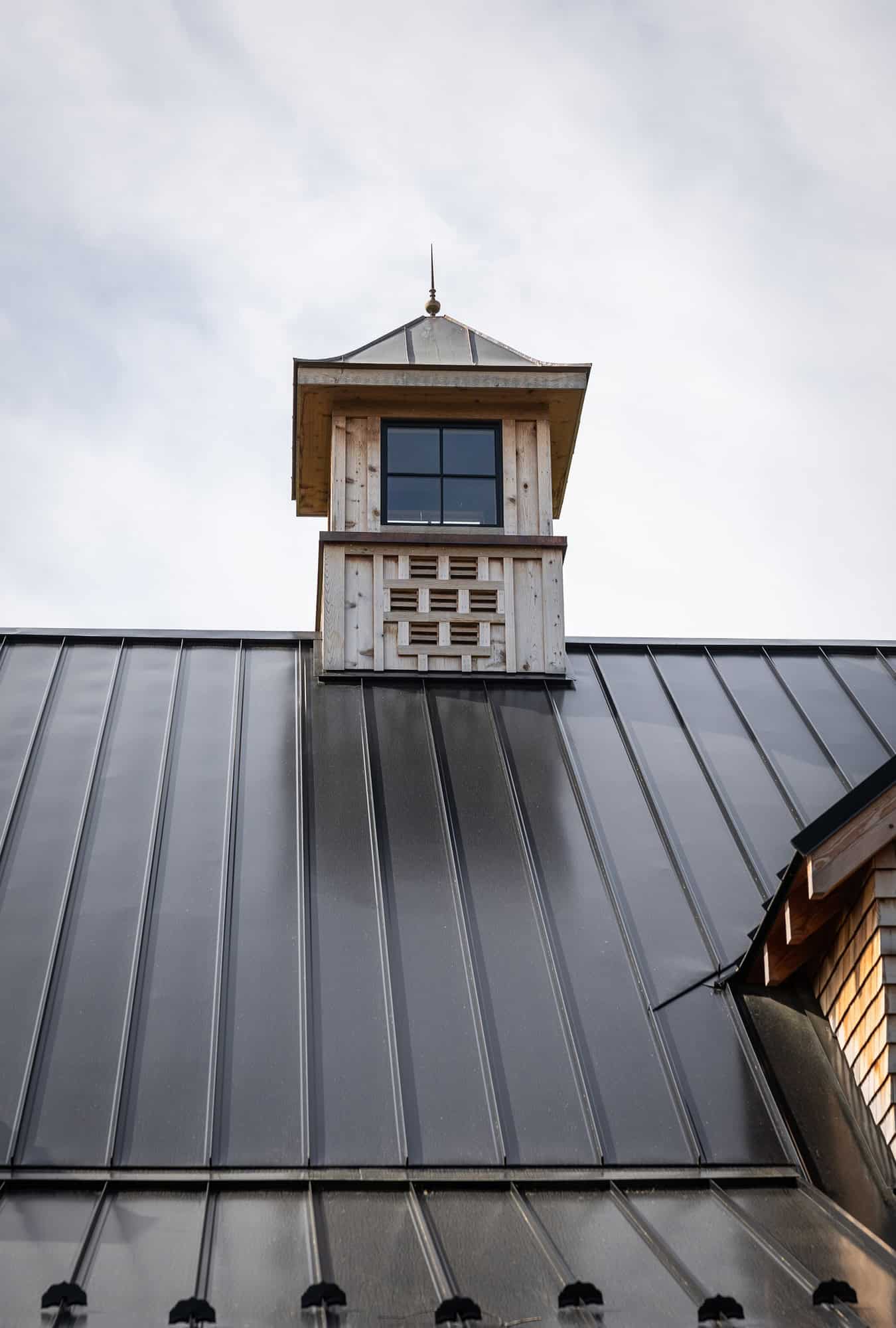 A close-up view of a metal roof with vertical seams on a luxury horse barn, featuring a small wooden cupola with a window and decorative latticework under a cloudy sky.