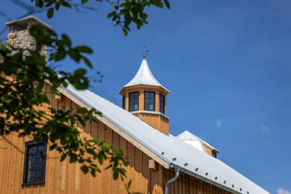 A wooden bank barn with a metal roof and a small cupola on top sits against a clear blue sky, while green leaves partially frame the top left corner of the image.