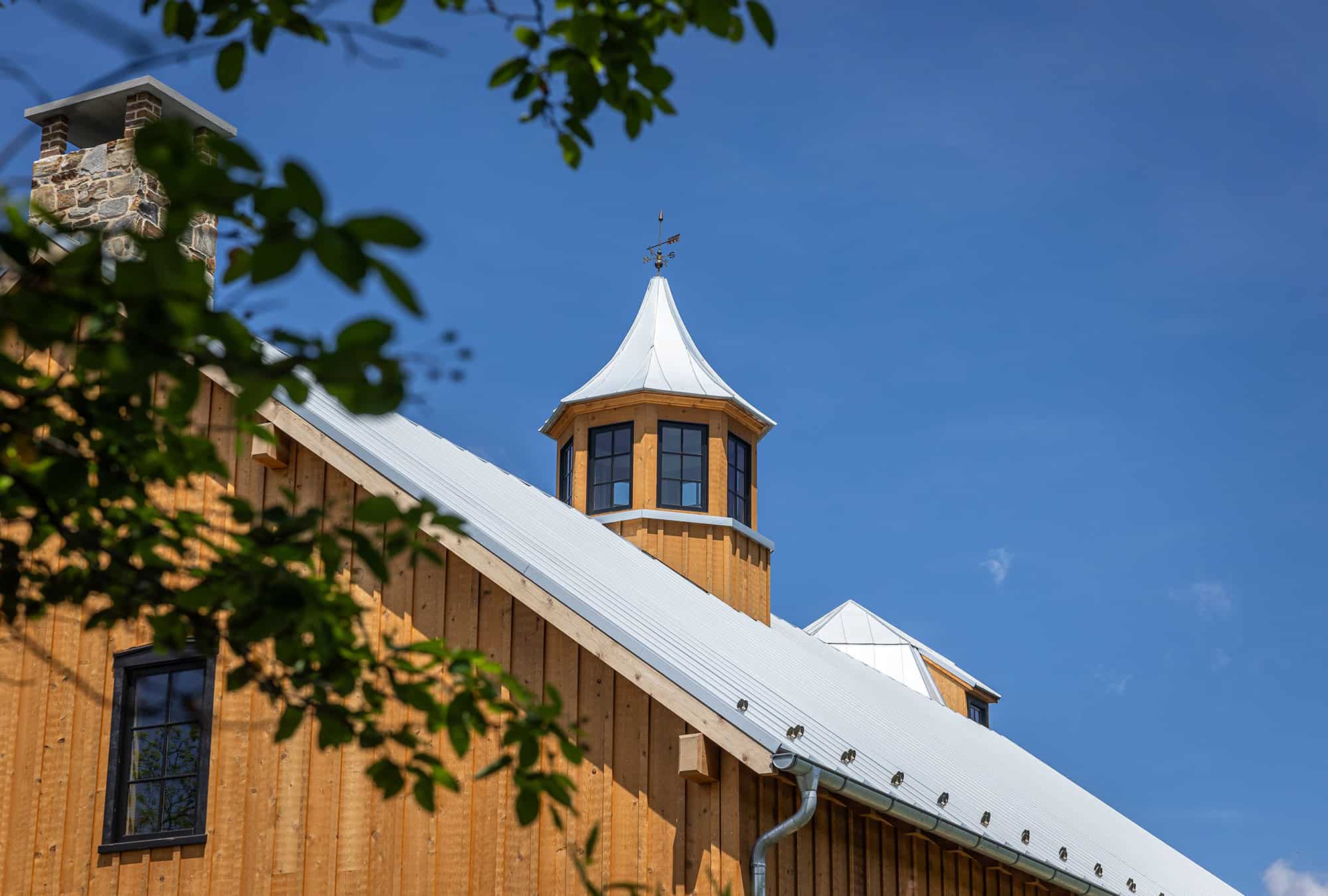 A wooden bank barn with a metal roof and a small cupola on top sits against a clear blue sky, while green leaves partially frame the top left corner of the image.