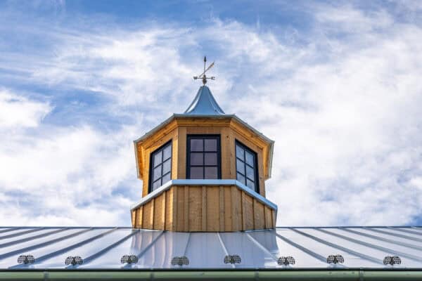 A wooden cupola with large windows and a weather vane sits atop the metal roof of a luxury horse barn, set against a blue sky with scattered clouds.