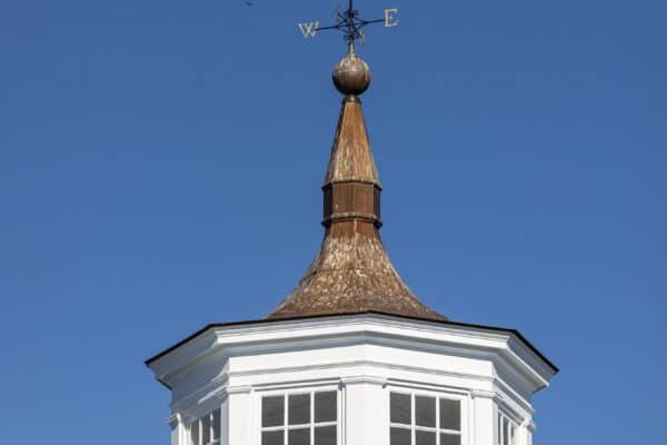 A white cupola with large windows sits atop a luxury horse barn, featuring a weather vane shaped like a horse and directional letters, all set against a clear blue sky.
