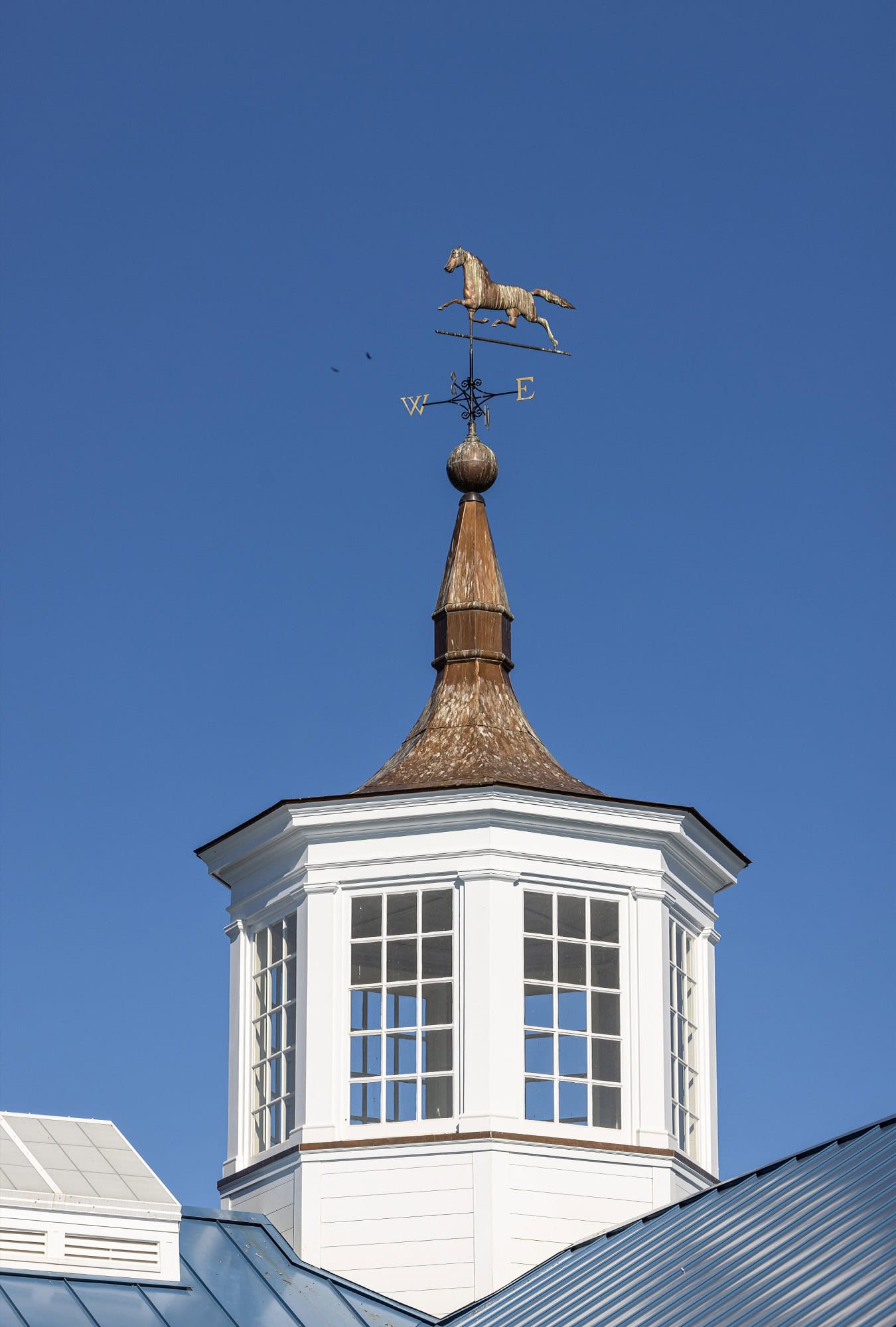 A white cupola with large windows sits atop a luxury horse barn, featuring a weather vane shaped like a horse and directional letters, all set against a clear blue sky.