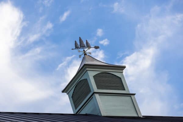 A weather vane shaped like a sailing ship sits atop a small, light green cupola with louvered vents, crowning a luxury horse barn beneath a blue sky with scattered white clouds.