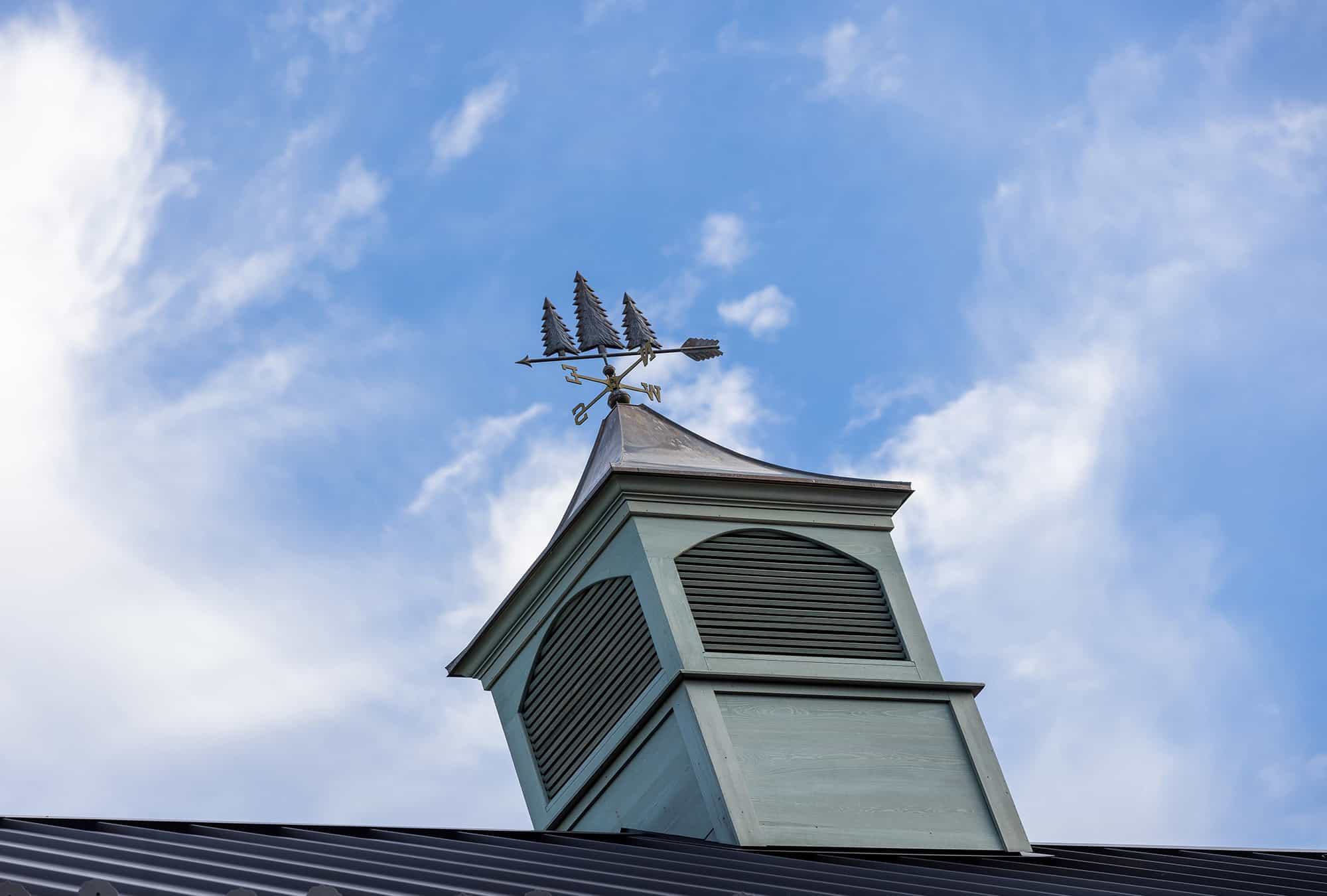 A weather vane shaped like a sailing ship sits atop a small, light green cupola with louvered vents, crowning a luxury horse barn beneath a blue sky with scattered white clouds.