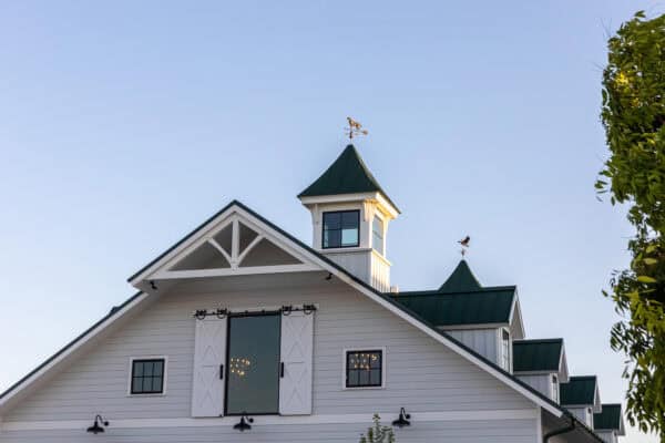White barn with green roofs and a central cupola, featuring classic barn doors, small windows, and weather vanes on top. This luxury horse barn sits under a clear blue sky, with part of a leafy tree visible on the right.