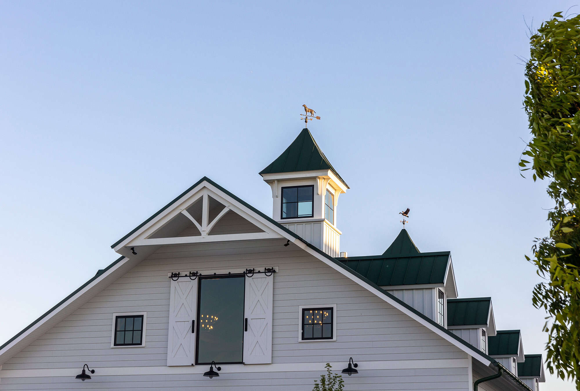 White barn with green roofs and a central cupola, featuring classic barn doors, small windows, and weather vanes on top. This luxury horse barn sits under a clear blue sky, with part of a leafy tree visible on the right.