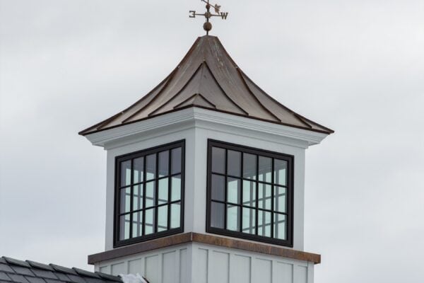 A cupola with large windows sits atop a slate roof of a luxury horse barn, featuring a weather vane with a bird silhouette pointing in different wind directions, set against a cloudy sky.