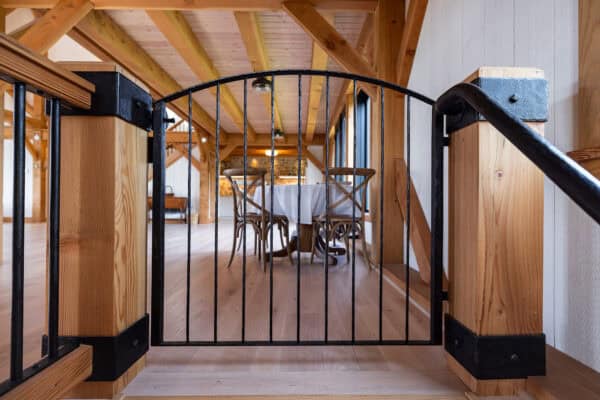 A black metal safety gate blocks the top of a wooden staircase, leading to a bright, rustic room with exposed wooden beams—evoking the charm of a renovated bank barn—and a table with several wooden chairs.