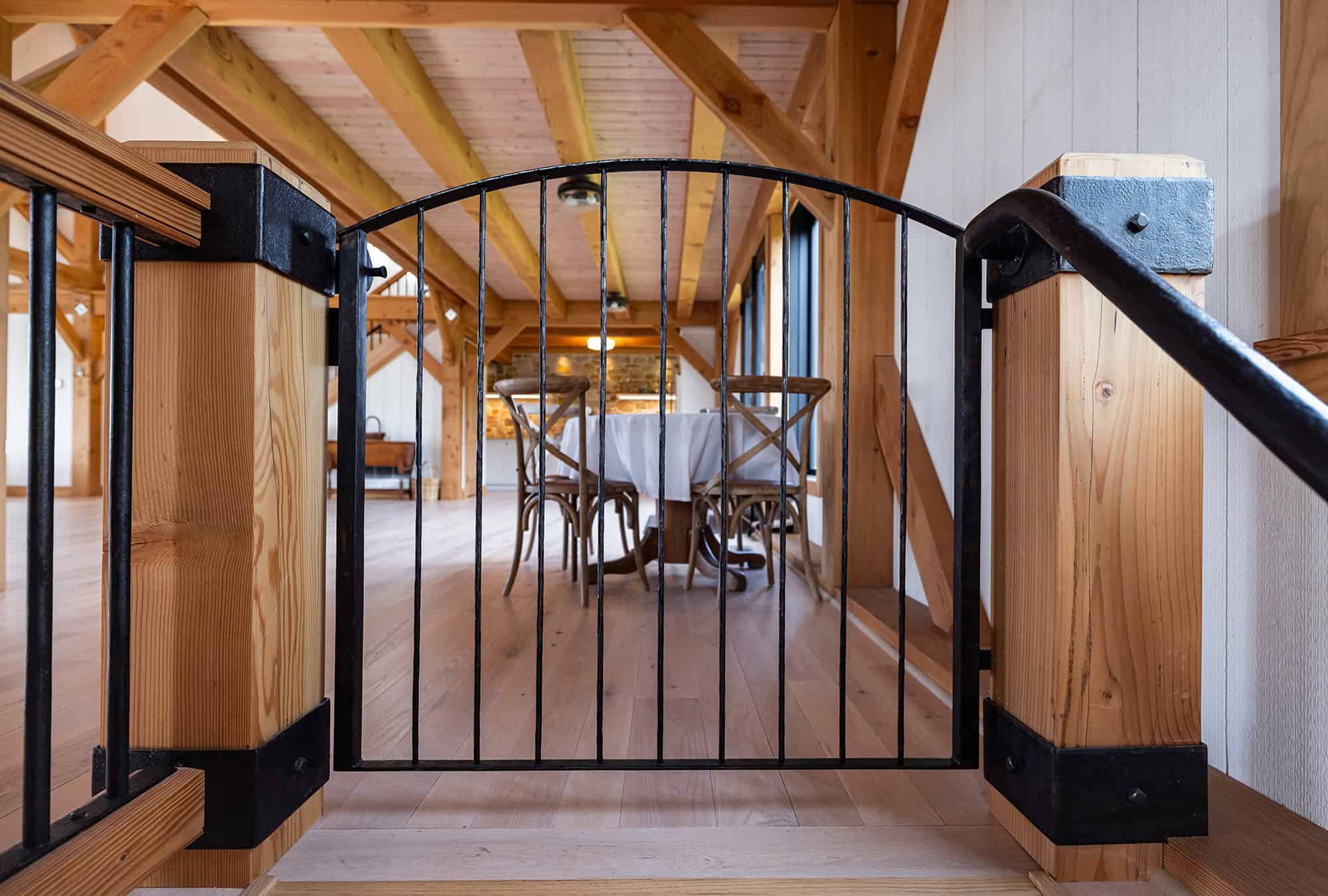 A black metal safety gate blocks the top of a wooden staircase, leading to a bright, rustic room with exposed wooden beams—evoking the charm of a renovated bank barn—and a table with several wooden chairs.