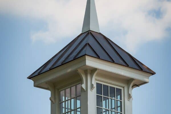 A white cupola with a pointed roof and large windows sits atop a shingled roof, hinting at a luxury home beneath, set against a blue sky with clouds and partially framed by tree leaves.