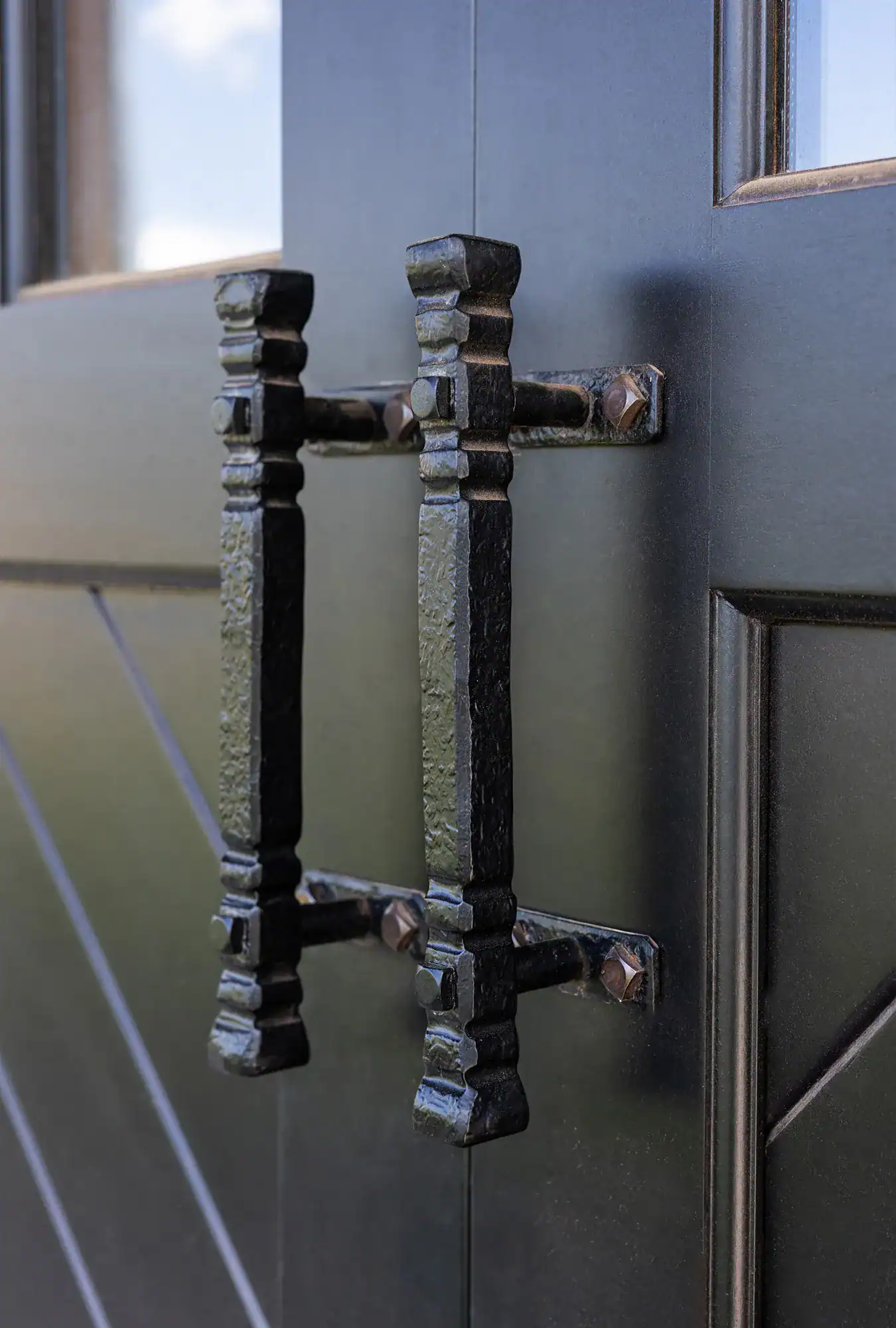 Close-up of two ornate, black metal door handles mounted on a dark wooden door with decorative paneling—perfect for adding a rustic touch to a luxury horse barn. The textured handles are attached with visible bolts for added charm.