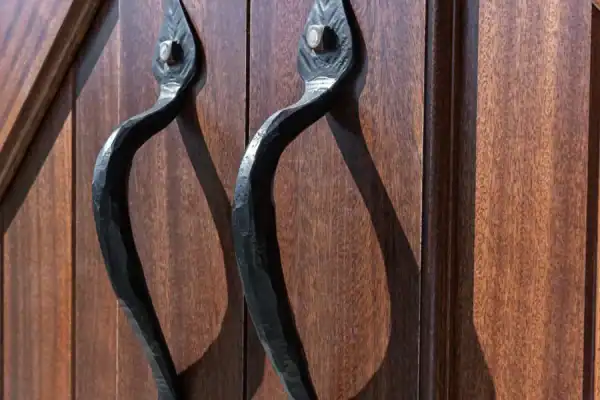 Close-up of two rustic, black metal handles on wooden double doors with a rich brown, textured finish—perfect for a luxury home or a renovated party barn. The handles have a curved, handcrafted design and are mounted vertically.