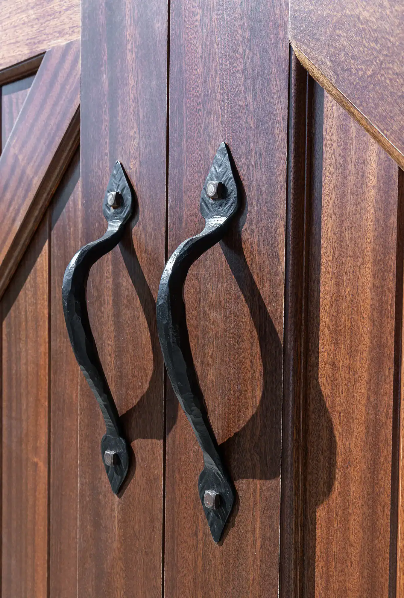 Close-up of two rustic, black metal handles on wooden double doors with a rich brown, textured finish—perfect for a luxury home or a renovated party barn. The handles have a curved, handcrafted design and are mounted vertically.