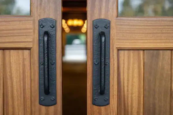 Close-up of two wooden doors slightly open, each with a long, black metal handle—hinting at the inviting warmth inside this luxury horse barn. Warm lights spill out, offering a glimpse of exquisite interiors beyond the doorway.