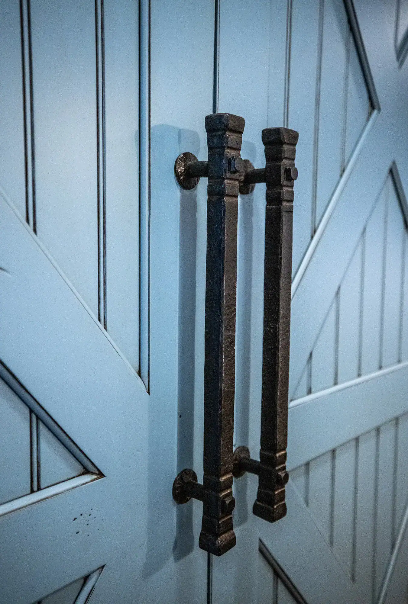 Close-up of two large, black metal door handles mounted vertically on a light blue, wooden barn-style door with diagonal and vertical panels—an elegant touch often seen in luxury home or bank barn designs.