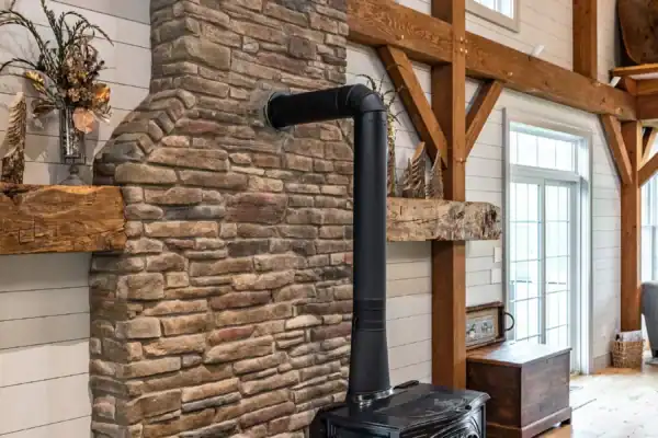 A black wood-burning stove with a tall chimney pipe sits on a raised hearth in front of a tall stone fireplace in a bright, rustic party barn living room with exposed wooden beams and large windows.