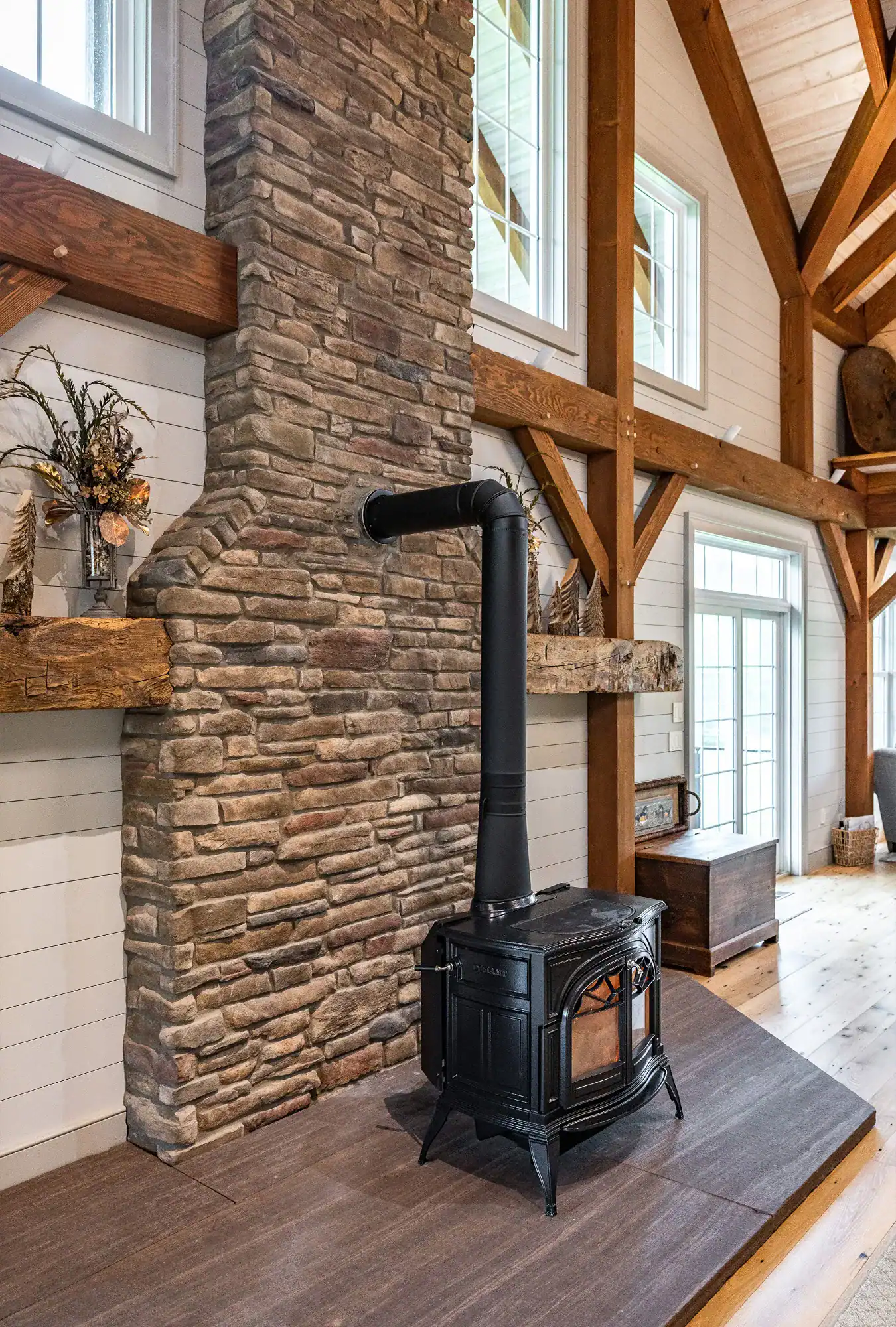 A black wood-burning stove with a tall chimney pipe sits on a raised hearth in front of a tall stone fireplace in a bright, rustic party barn living room with exposed wooden beams and large windows.