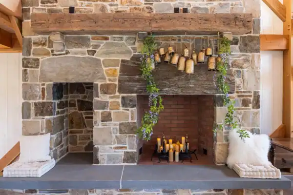A rustic stone fireplace with a wooden mantel, hanging brass bells, green foliage, and grouped candles inside creates the perfect focal point for a cozy party barn. Two cushioned seats sit on either side of the fireplace.