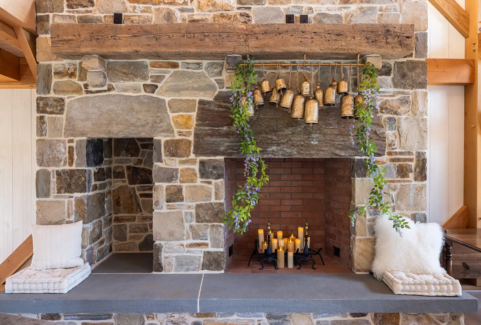 A rustic stone fireplace with a wooden mantel, hanging brass bells, green foliage, and grouped candles inside creates the perfect focal point for a cozy party barn. Two cushioned seats sit on either side of the fireplace.