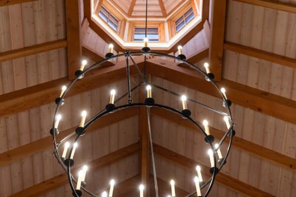 A large circular chandelier with lit candles hangs from a high, wooden, vaulted ceiling with exposed beams and an octagonal skylight above, adding rustic charm to this luxury home’s party barn.