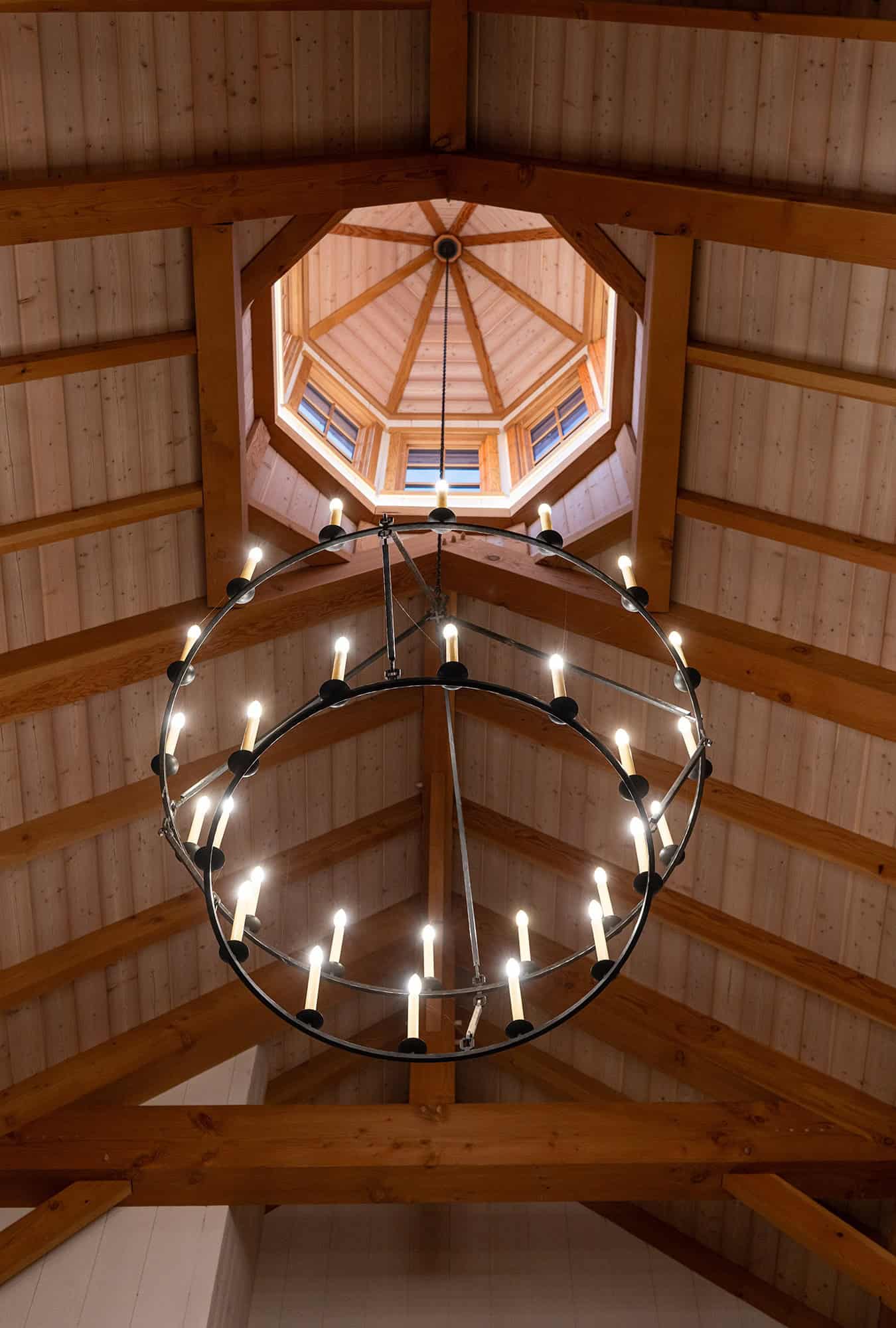 A large circular chandelier with lit candles hangs from a high, wooden, vaulted ceiling with exposed beams and an octagonal skylight above, adding rustic charm to this luxury home’s party barn.