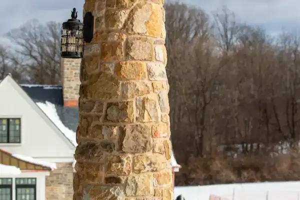 A sturdy stone column supports a wooden overhang on a snowy porch of this luxury home, with a black lantern attached. In the background, there is a house, leafless trees, and snow-covered ground.