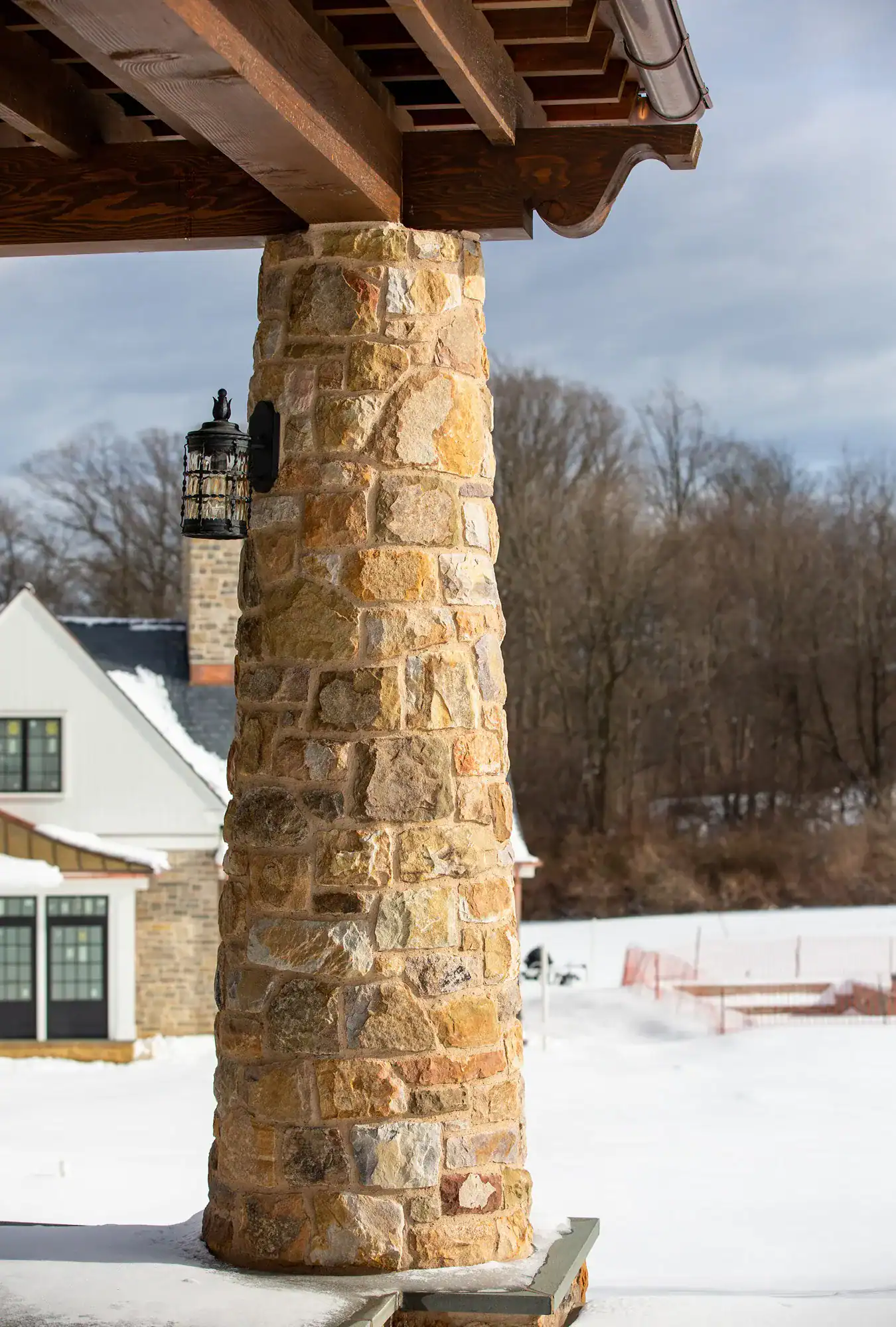 A sturdy stone column supports a wooden overhang on a snowy porch of this luxury home, with a black lantern attached. In the background, there is a house, leafless trees, and snow-covered ground.