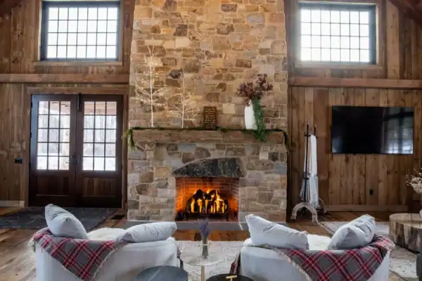A cozy rustic living room, inspired by a luxury home, features a tall stone fireplace, lit fire, two curved gray sofas with plaid blankets, round side tables, wood-paneled walls, large windows, and a vaulted ceiling with a circular chandelier.
