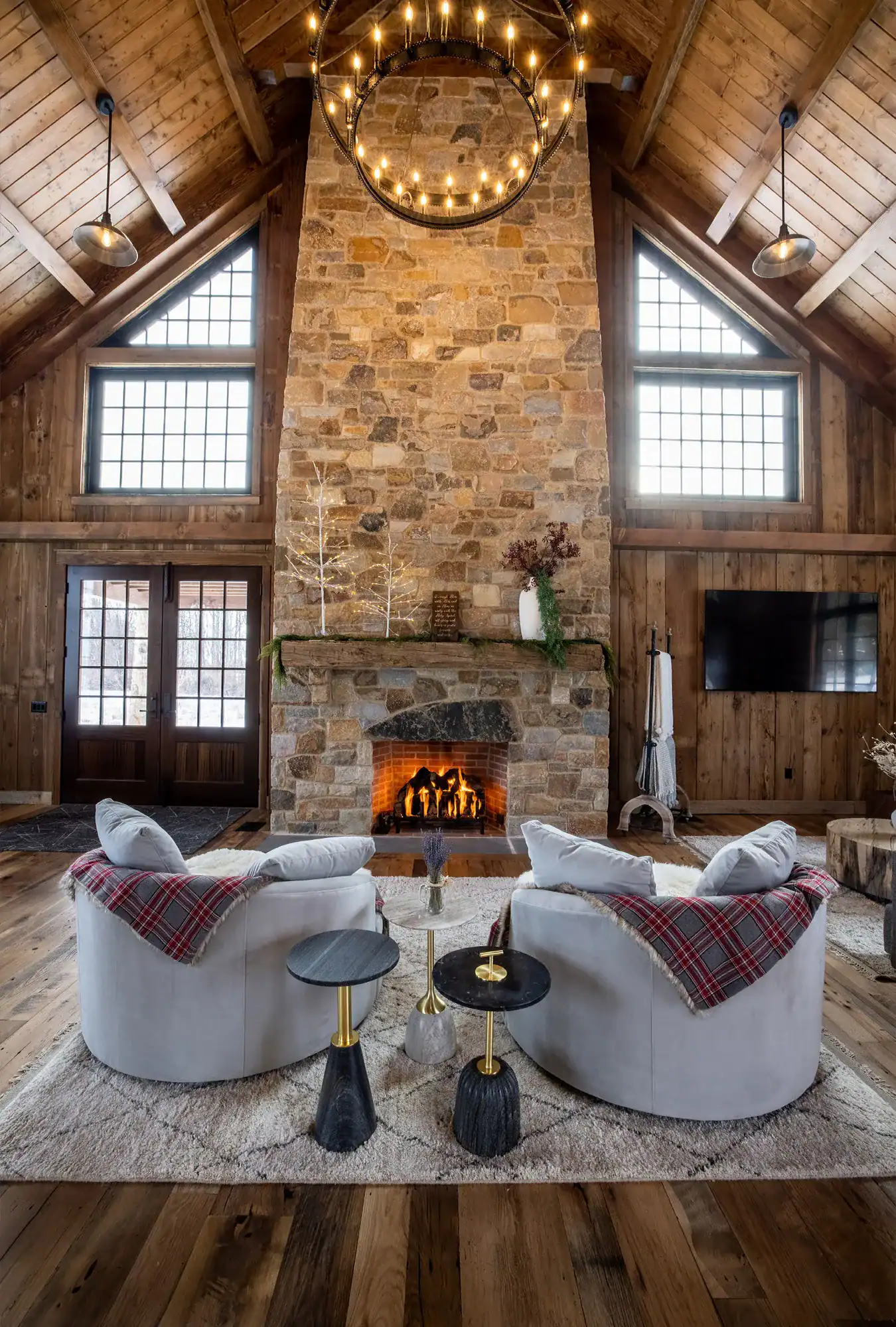 A cozy rustic living room, inspired by a luxury home, features a tall stone fireplace, lit fire, two curved gray sofas with plaid blankets, round side tables, wood-paneled walls, large windows, and a vaulted ceiling with a circular chandelier.