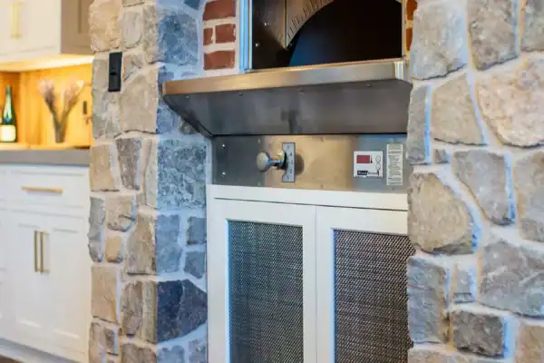 A built-in pizza oven with a metal front is set into a rustic stone wall in a luxury home kitchen with wood floors and white cabinetry. The oven features digital controls and a brick interior.