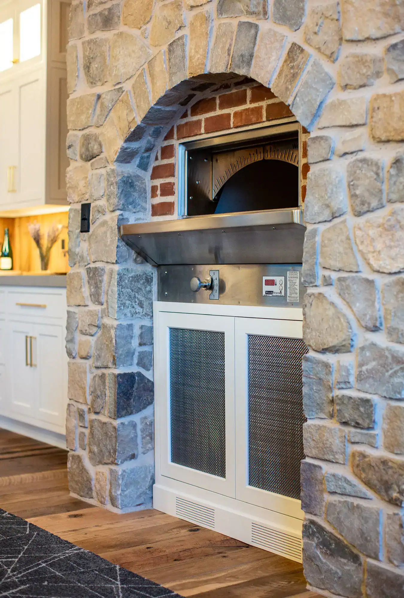 A built-in pizza oven with a metal front is set into a rustic stone wall in a luxury home kitchen with wood floors and white cabinetry. The oven features digital controls and a brick interior.