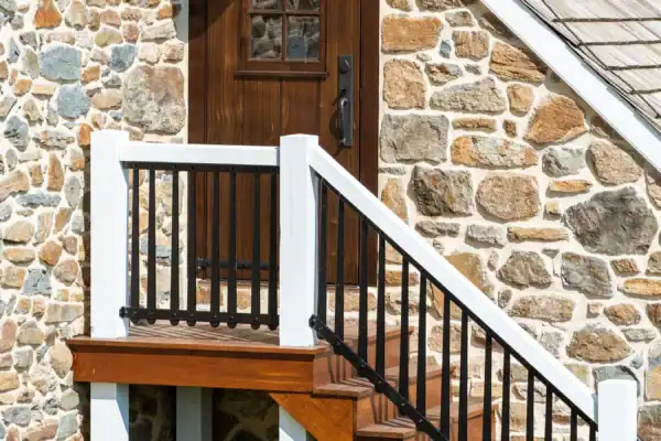 A wooden door with a small window is set in a stone wall, accessed by a stairway with black railings and white posts, and topped with an outdoor light fixture—classic features of a charming bank barn. Grass grows at the base of the structure.