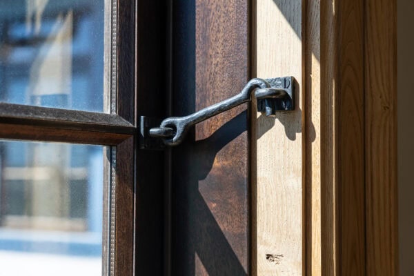A close-up of a window secured with a metal latch hook fastened to a wooden frame in a luxury horse barn. Sunlight streams through the glass, casting elegant shadows on the wood.